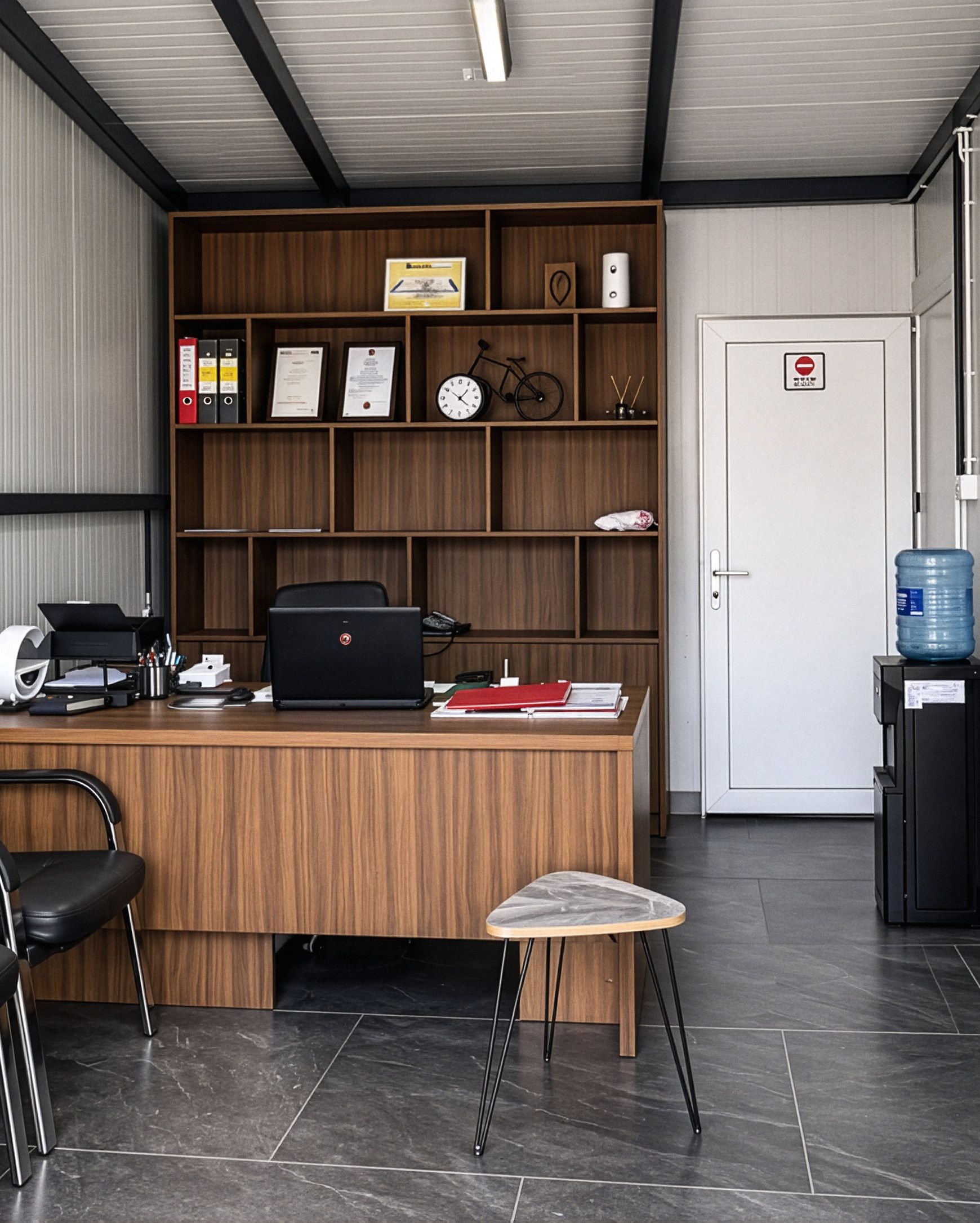 Das Büro: Schreibtisch, Holzregal, Besucherstühle und eine Innentür unter einer Decke mit Stahlträgern.